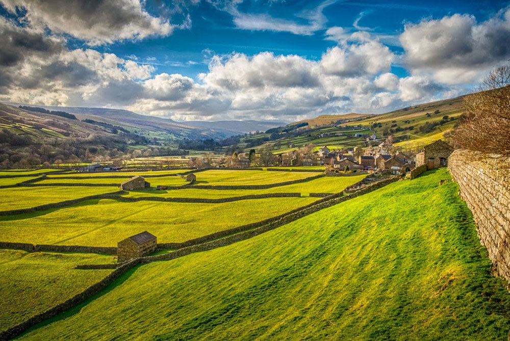 A photo of green fields with stone walls with a blue sky and clouds above.