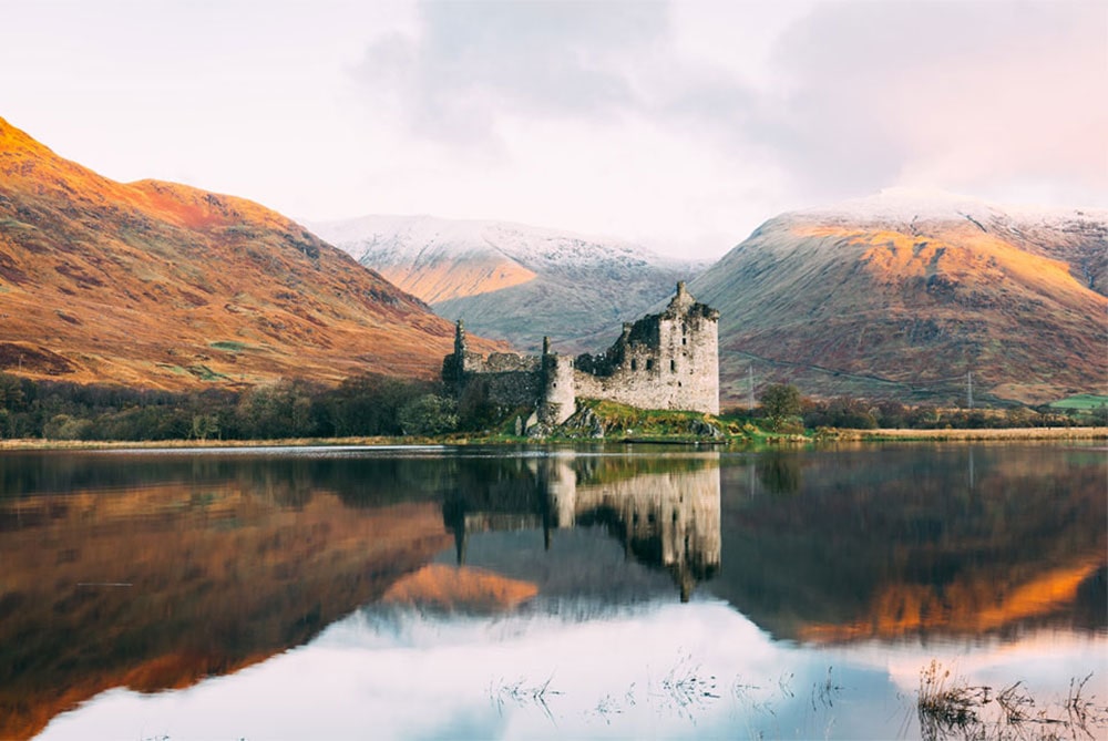 Ruins of Kilchurn Castle reflecting in the calm waters of Loch Awe, surrounded by autumn-colored hills and snow-capped mountains in the Scottish Highlands.